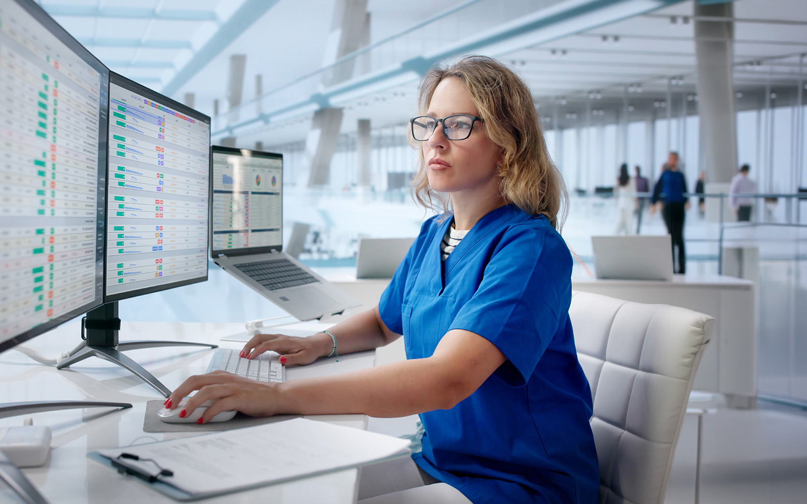 Healthcare professional in blue scrubs reviews data dashboards across multiple computer screens.