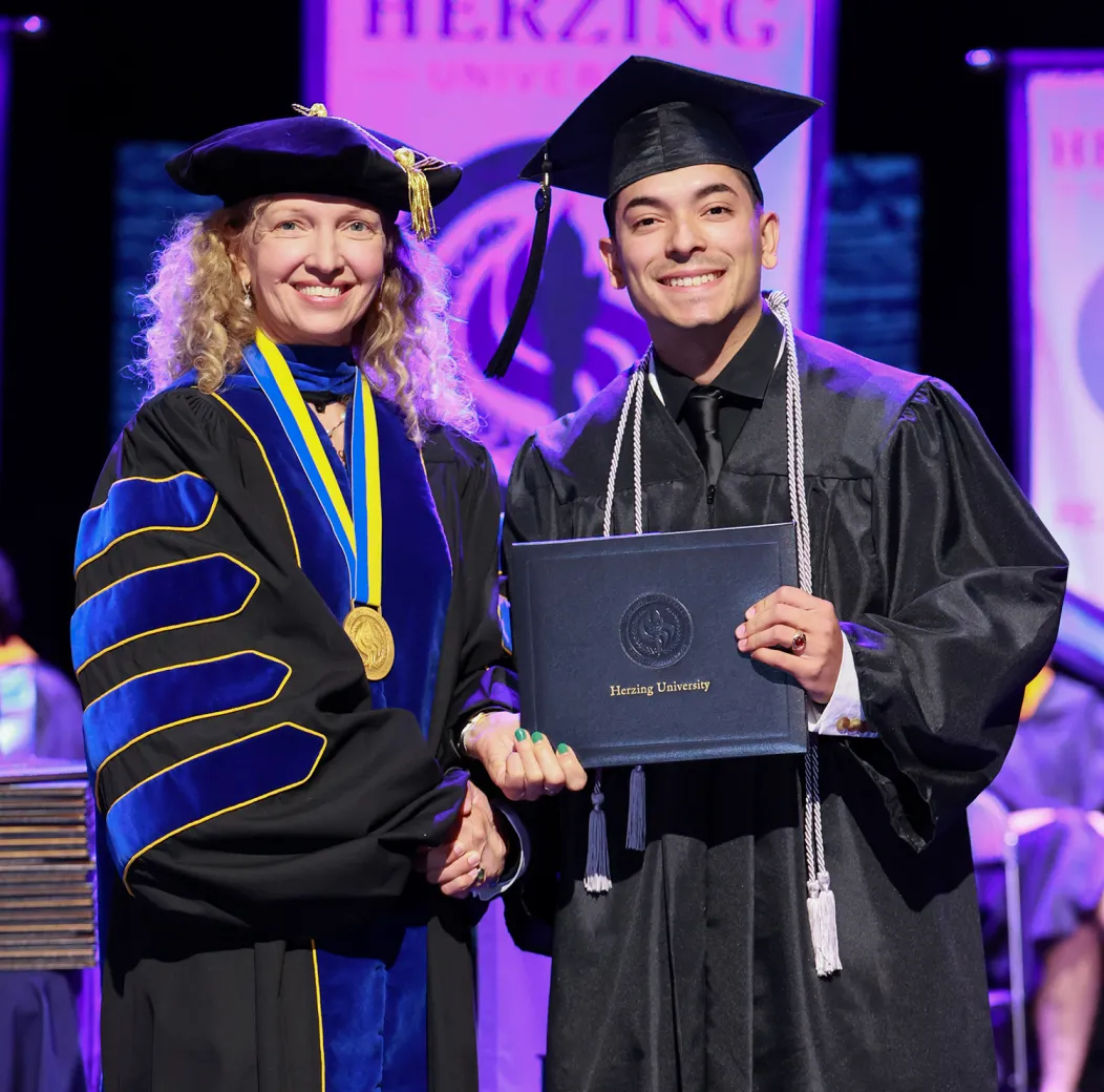 A proud graduate in a black cap and gown holding a Herzing University diploma, shaking hands with a smiling faculty member in academic regalia during a graduation ceremony, with university banners in the background