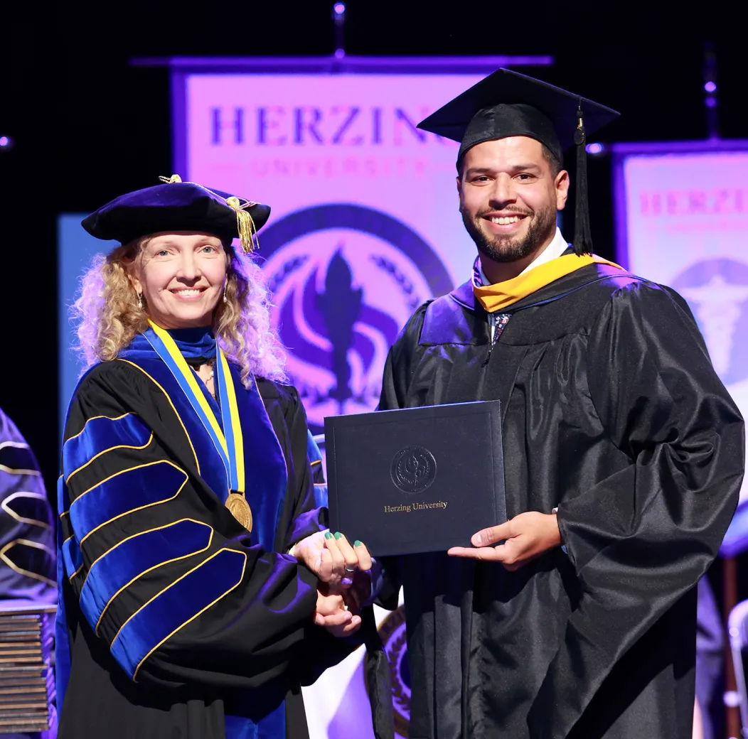 A graduate student wearing a black cap and gown with a gold hood holds a Herzing University diploma while shaking hands with university president in academic regalia during a formal graduation ceremony, with university banners displayed in the background.