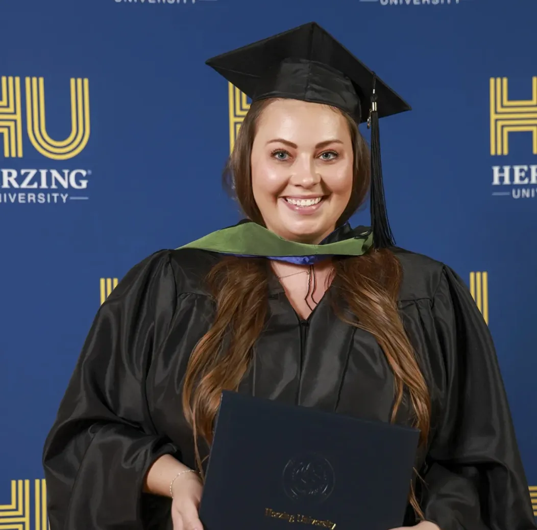 Graduate in cap and gown smiling and holding diploma in front of Herzing University backdrop