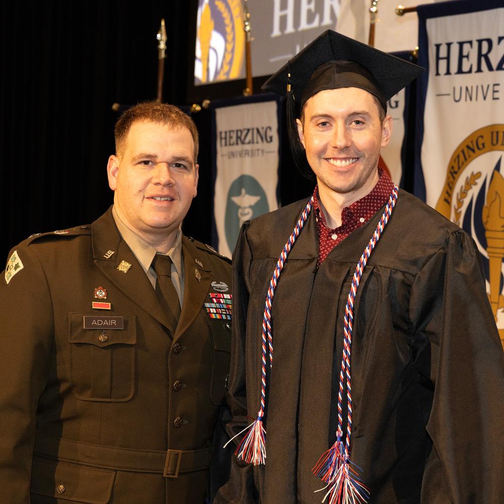 Herzing University graduate in cap and gown posing with a military service member in uniform during a graduation ceremony.