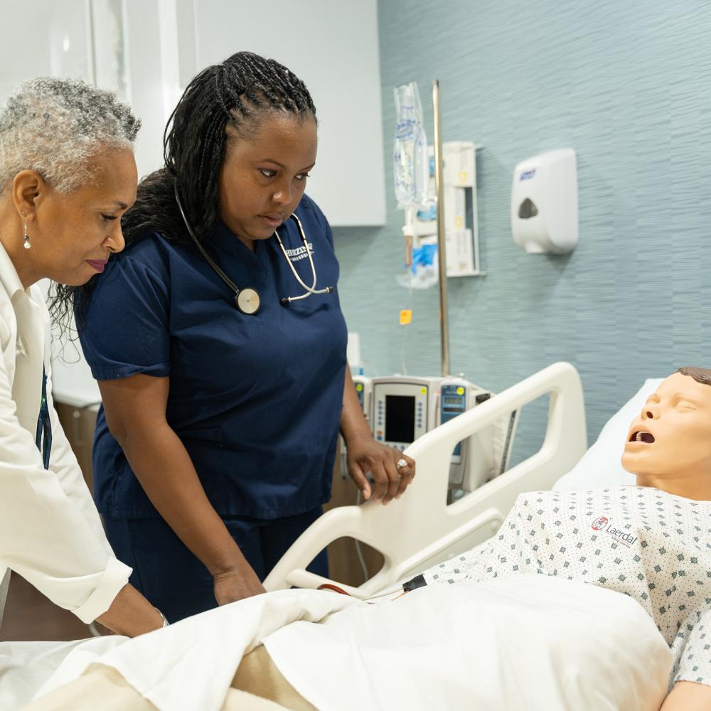 Herzing University nursing faculty member guiding a student through a hands-on patient care simulation with a medical training mannequin in a clinical lab.