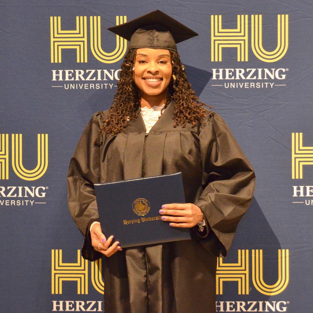 Smiling Herzing University graduate in a cap and gown holding a diploma in front of a branded backdrop at the graduation ceremony