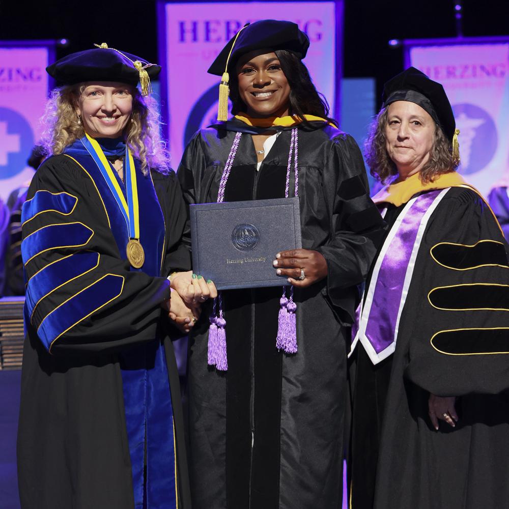 Herzing University DNP graduate in academic regalia holding a diploma and smiling while posing with two faculty members during a commencement ceremony.